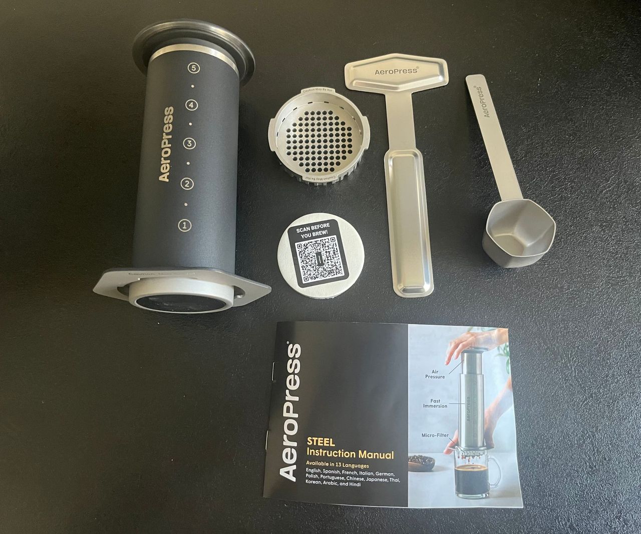 The Stainless Steel AeroPress and all of it's accessories laid out on a black kitchen counter.
