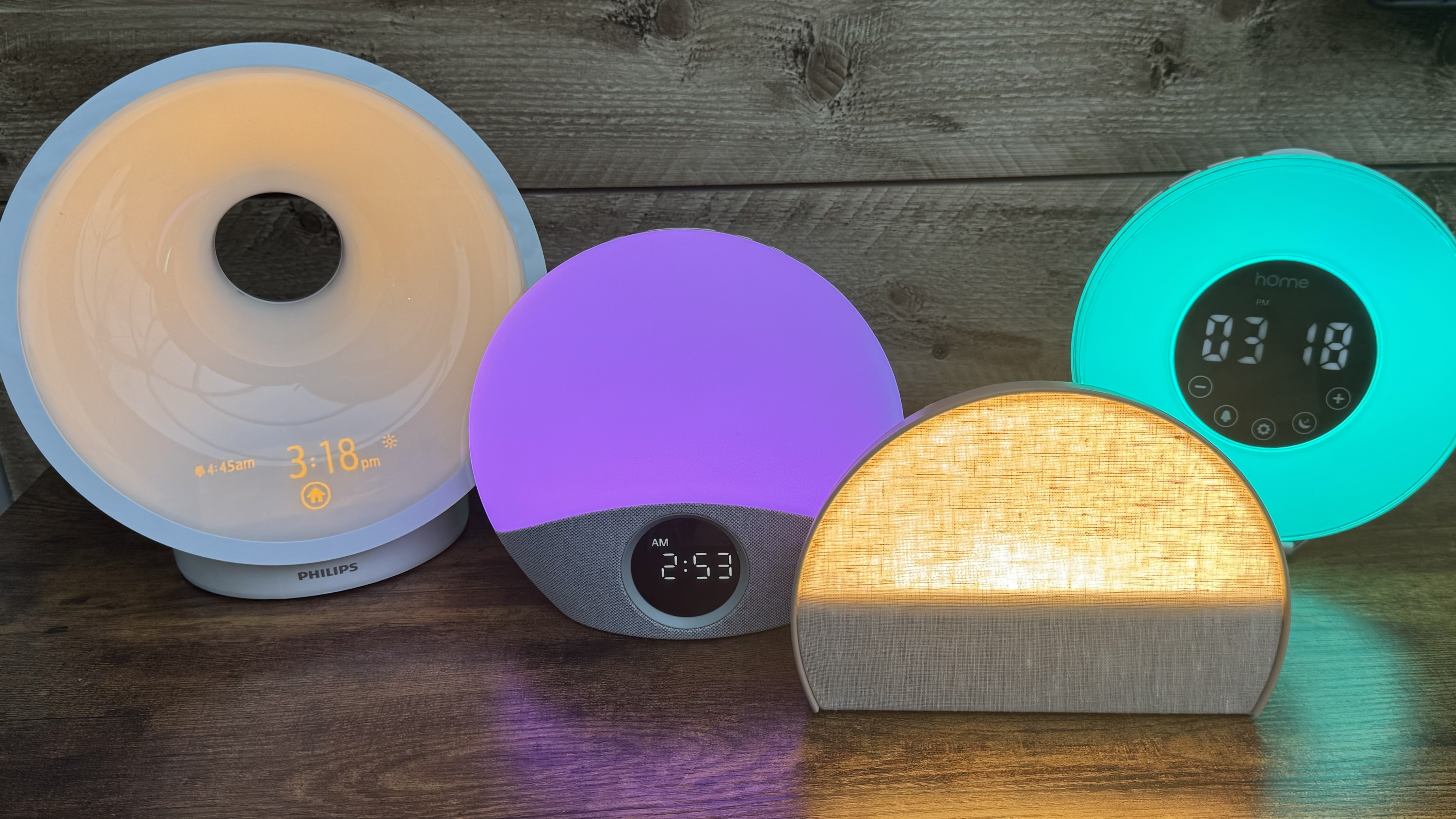 Four sunrise alarm clocks Black Friday deals with different coloured lights on - amber sunlight, purple light, blue light - on wooden surface.