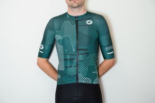 A white man in an emerald green cycling jersey and black cycling shorts stands against a white wall