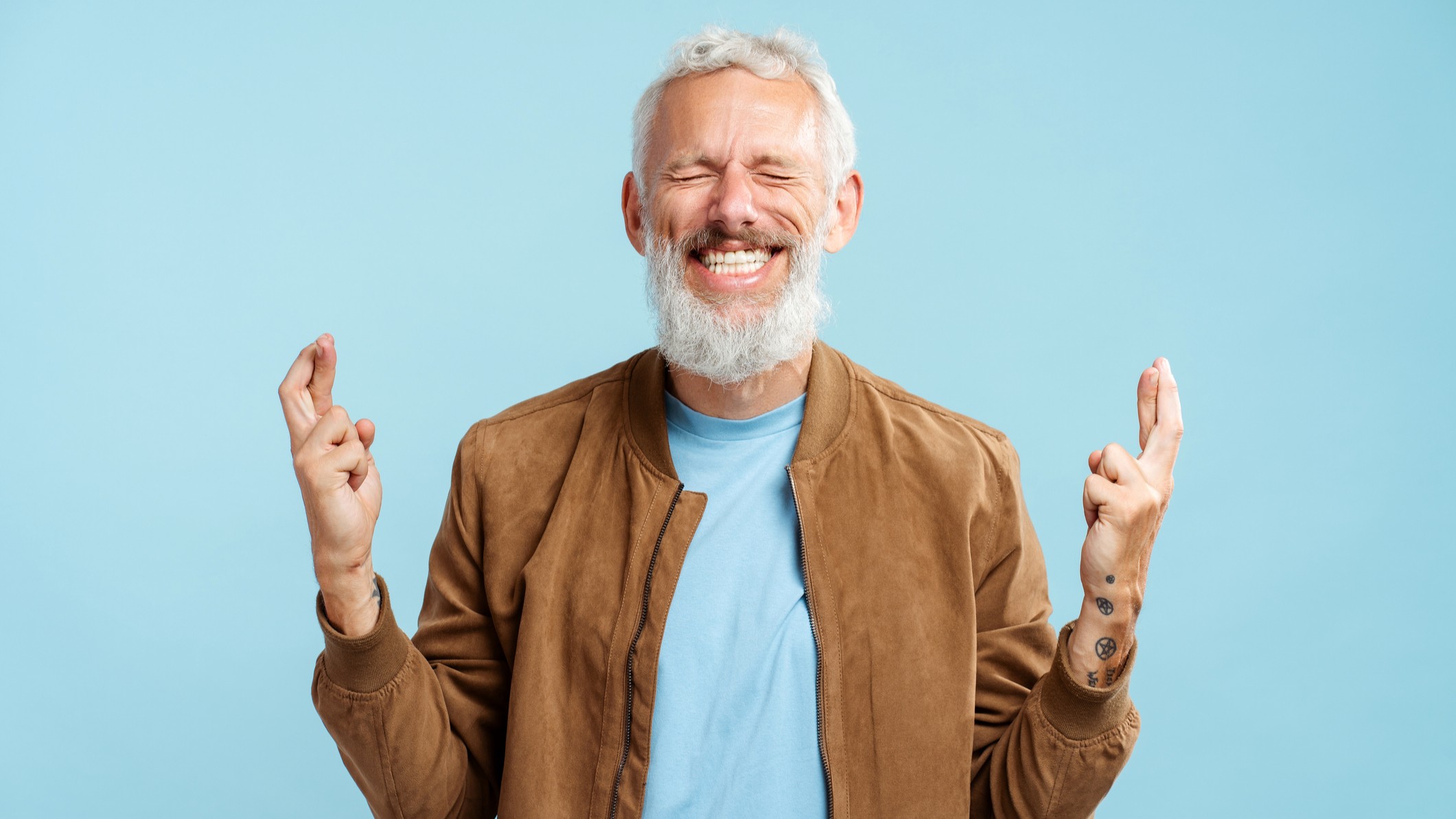 Portrait of smiling gray haired bearded mature man wearing stylish casual jacket with fingers crossed and eyes closed standing isolated on blue background. Concept of hope