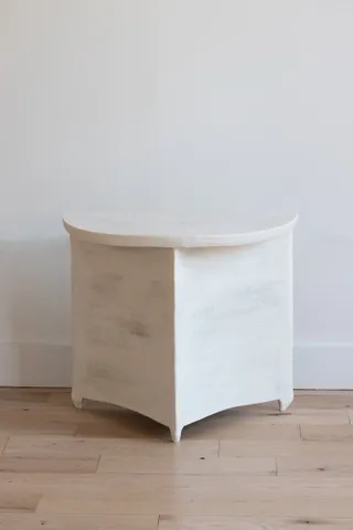 A wooden coffee table shaped as a half moon stands against a white background referencing its monochromatic tones on a wooden floor.