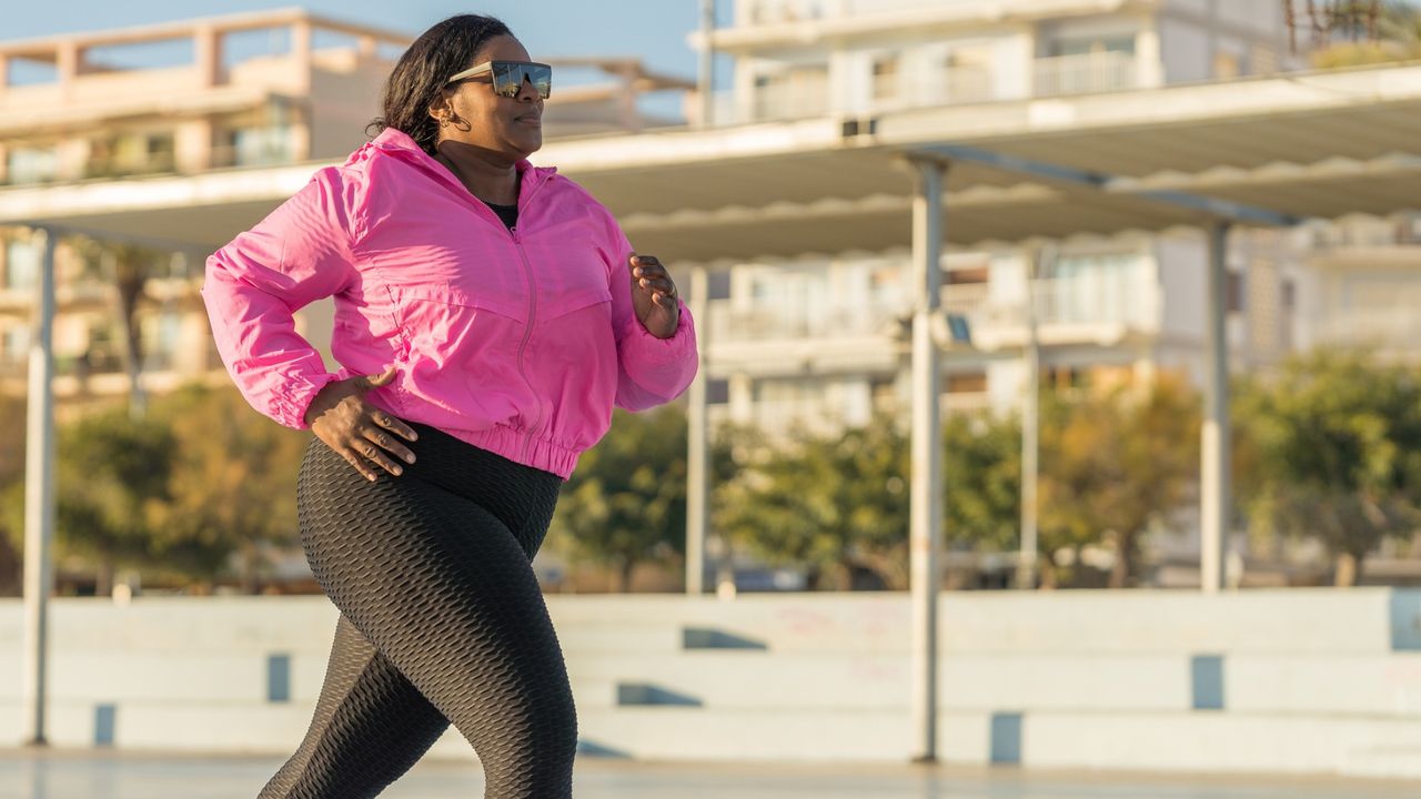 woman wearing a bright pink running jacket, black leggings and sunglasses running outside with a sunny residential background