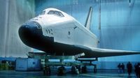 space shuttle in a large hangar with people walking underneath the wing on the right. 