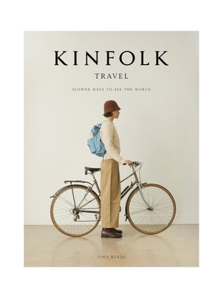 Kinfolk Travel: Slower Ways to See the World Book in beige features a woman standing next to her bike dressed in sandy clothes and wearing a blue backpack.