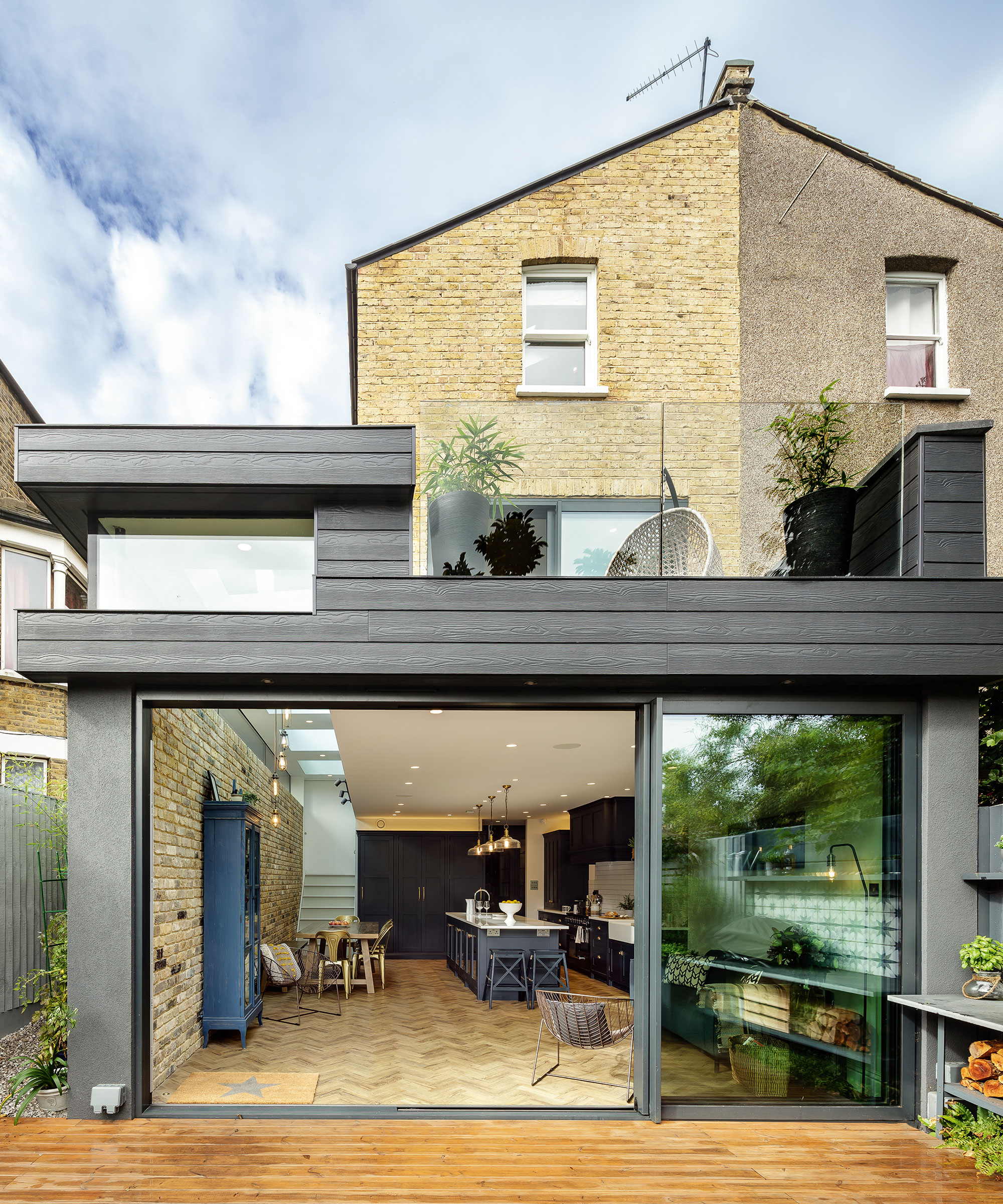 semi-detached house with large rear kitchen extension featuring sliding glass doors leading out to a decked patio. It has dark external wood cladding and a first floor terrace on top of the flat roof