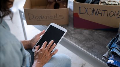 A woman looks at a tablet while sitting by cardboard boxes marked with the word donations.
