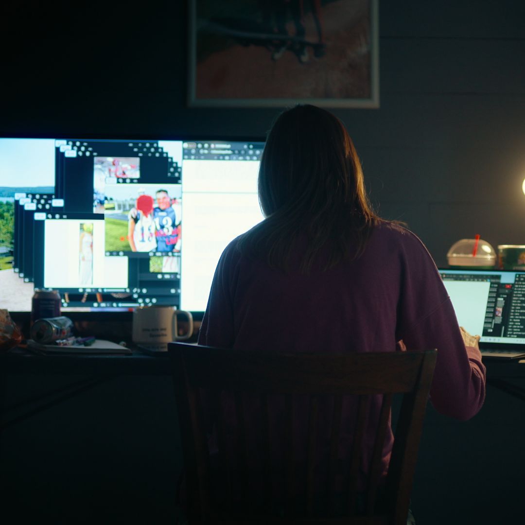 A woman facing away from the camera works on a desktop computer and laptop in a dark room with a desk lamp on, in &#039;Unknown Number: The High School Catfish&#039; on Netflix.