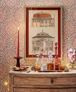 Antique oak chest of drawers with sparkling crystalware and antique glass, a lit red candle and a drinks tray with an architectural framed print and patterned wallpaper.