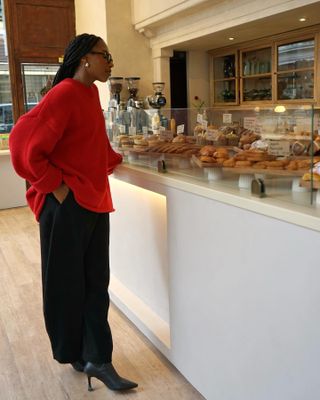 British fashion style influencers Taffy Msipa looking at pastries at a cafe wearing a chic outfit with an oversize red sweater, black trousers, and heeled boots