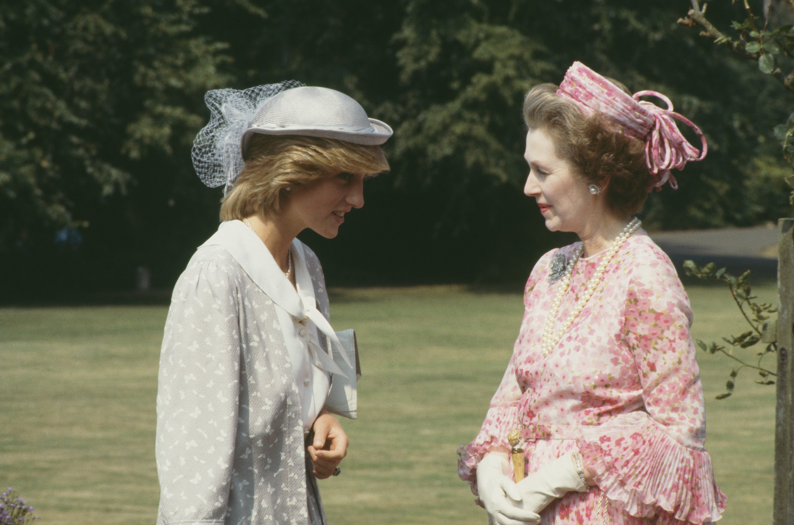 Princess Diana talking to Raine Spencer in front of some trees, both wearing dresses and hats