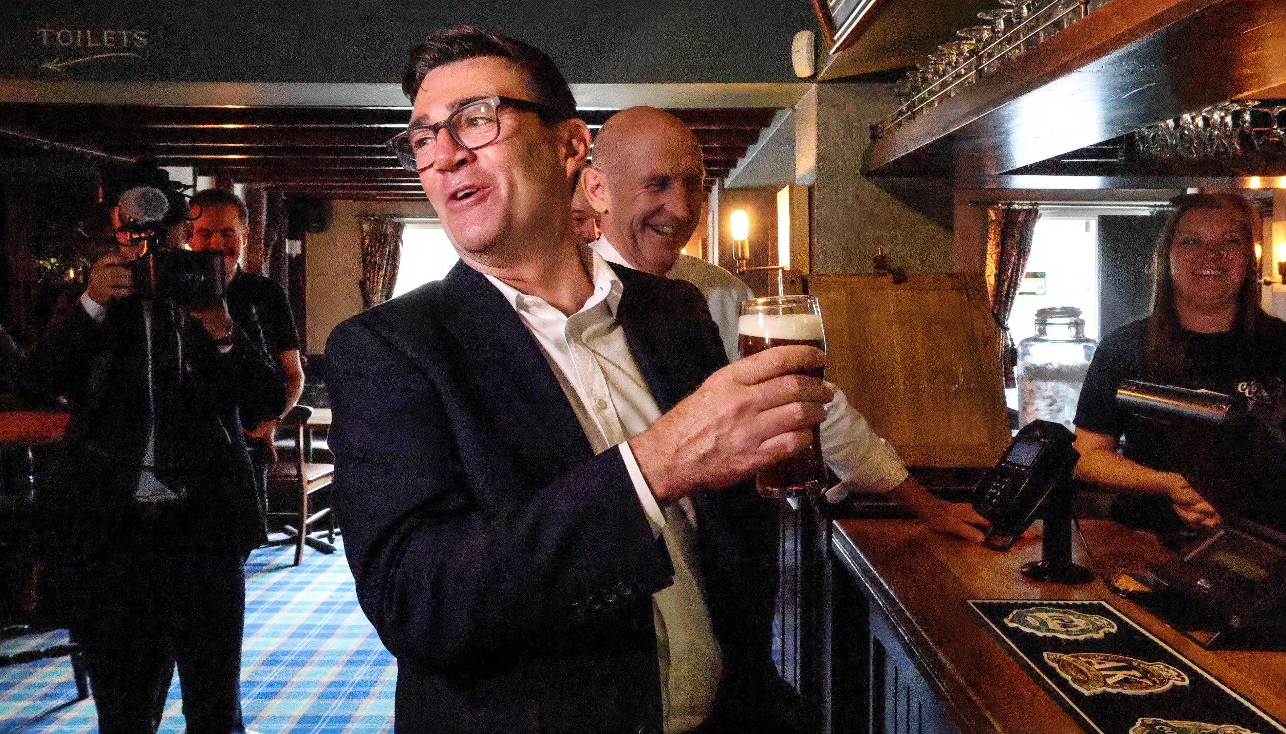 Prime Minister Andy Burnham reacts while holding a pint of beer as he visits the Hare Inn in Harlow, Essex with the Chancellor John Healy (C) on July 23, 2026 in Harlow, United Kingdom. Prime Minister Andy Burnham has announced a 20% cut in business rates for 32,000 hospitality businesses including pubs, clubs, and live music venues in England, which will take effect from April 2027