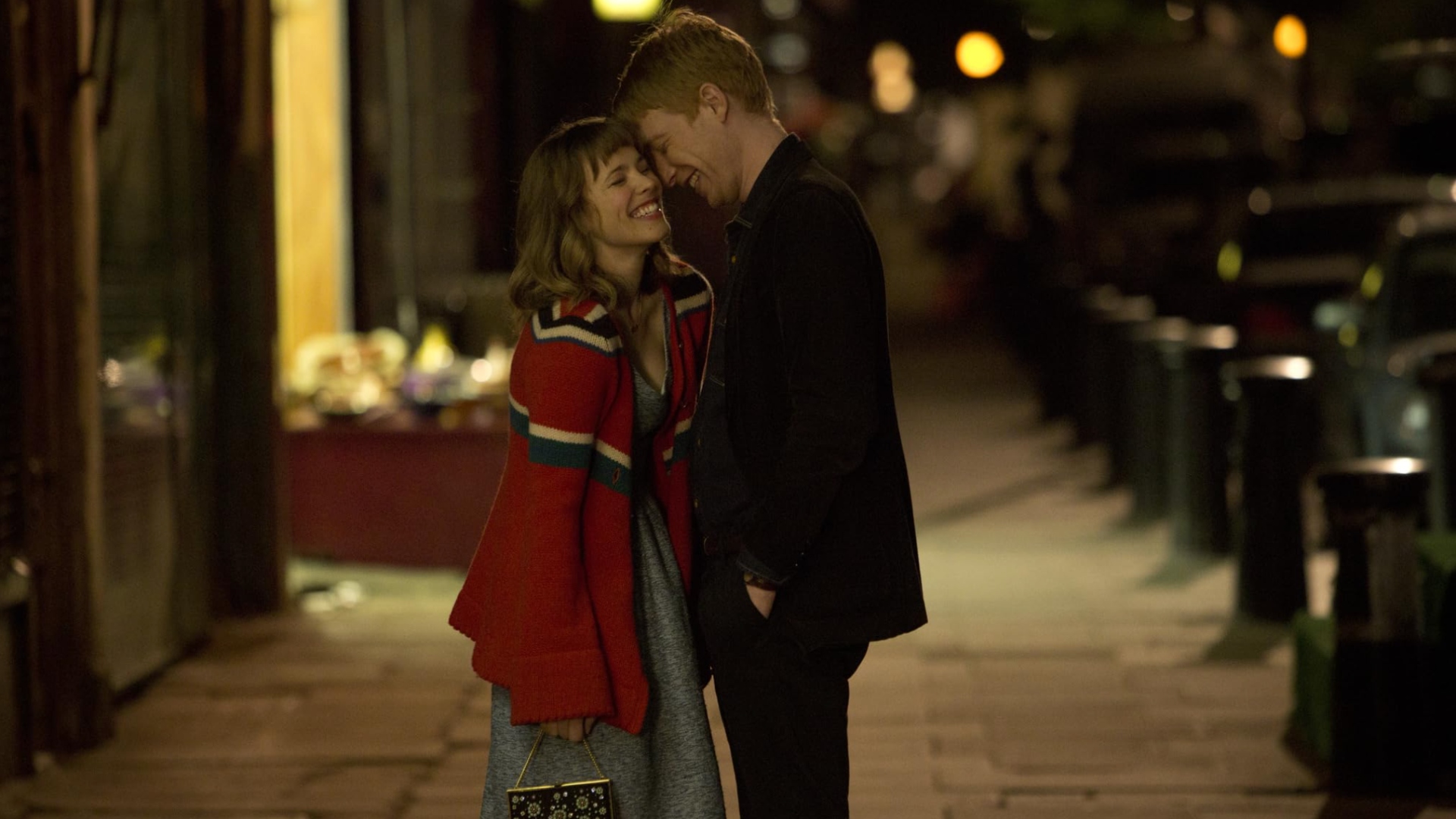 A man in a black coat and a woman in a blue dress and red jacket, stood together on the sidewalk, pressing their heads together lovingly.