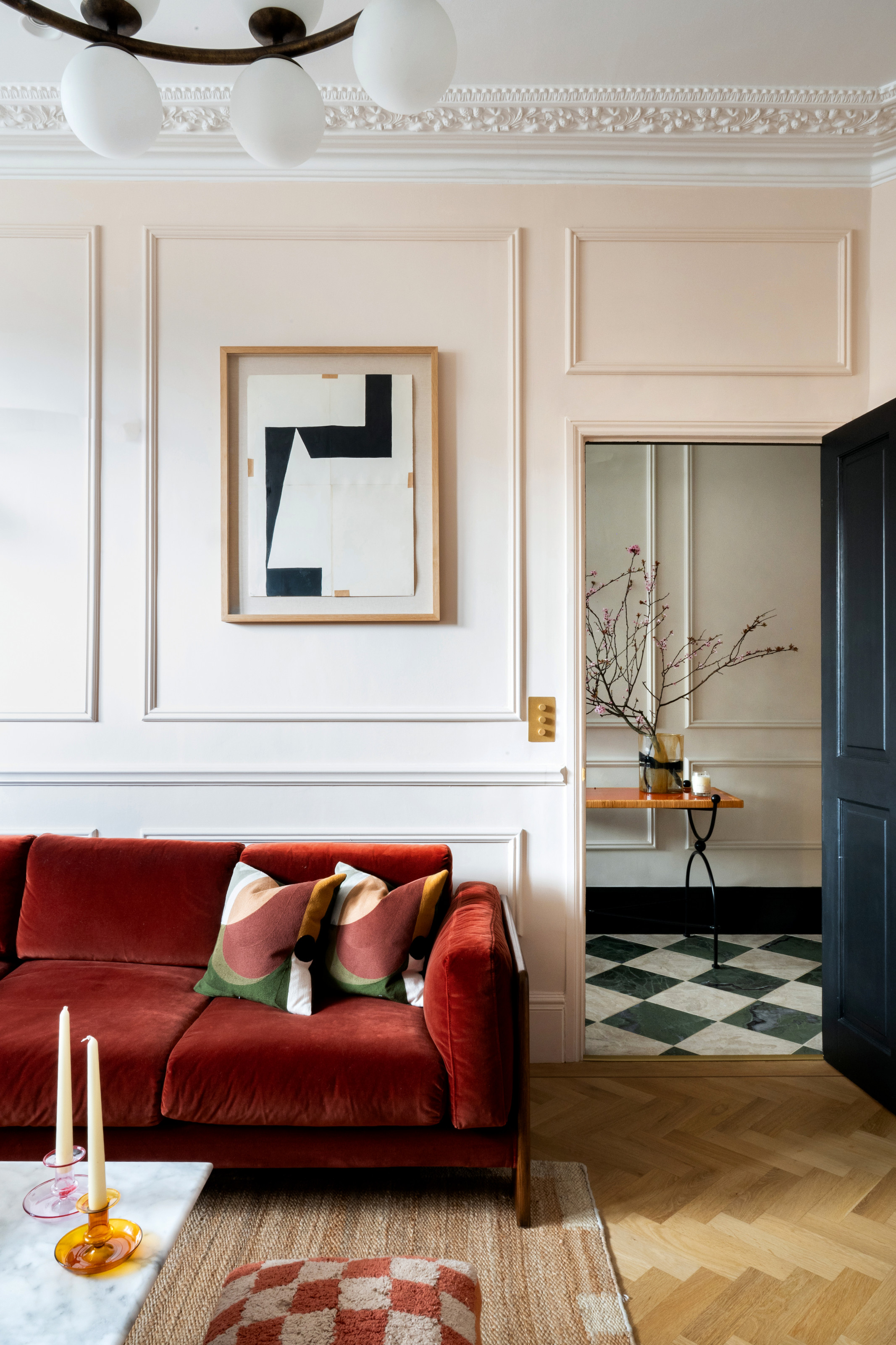 A living room with white panelled walls, burnt red sofa, abstract black and white artwork in a pale wood frame, pale wood herringbone flooring and abstract red, white and green cushions