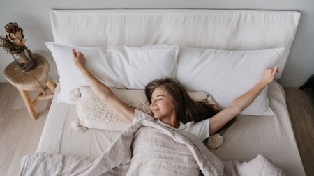Woman wakes smiling and stretching in a bed with light-coloured bedding.