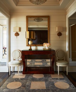 make your home feel like a sanctuary, classic entryway with symmetry, tiled floor, cream walls, mahogany console, matching chairs each side, mirror, wall lights