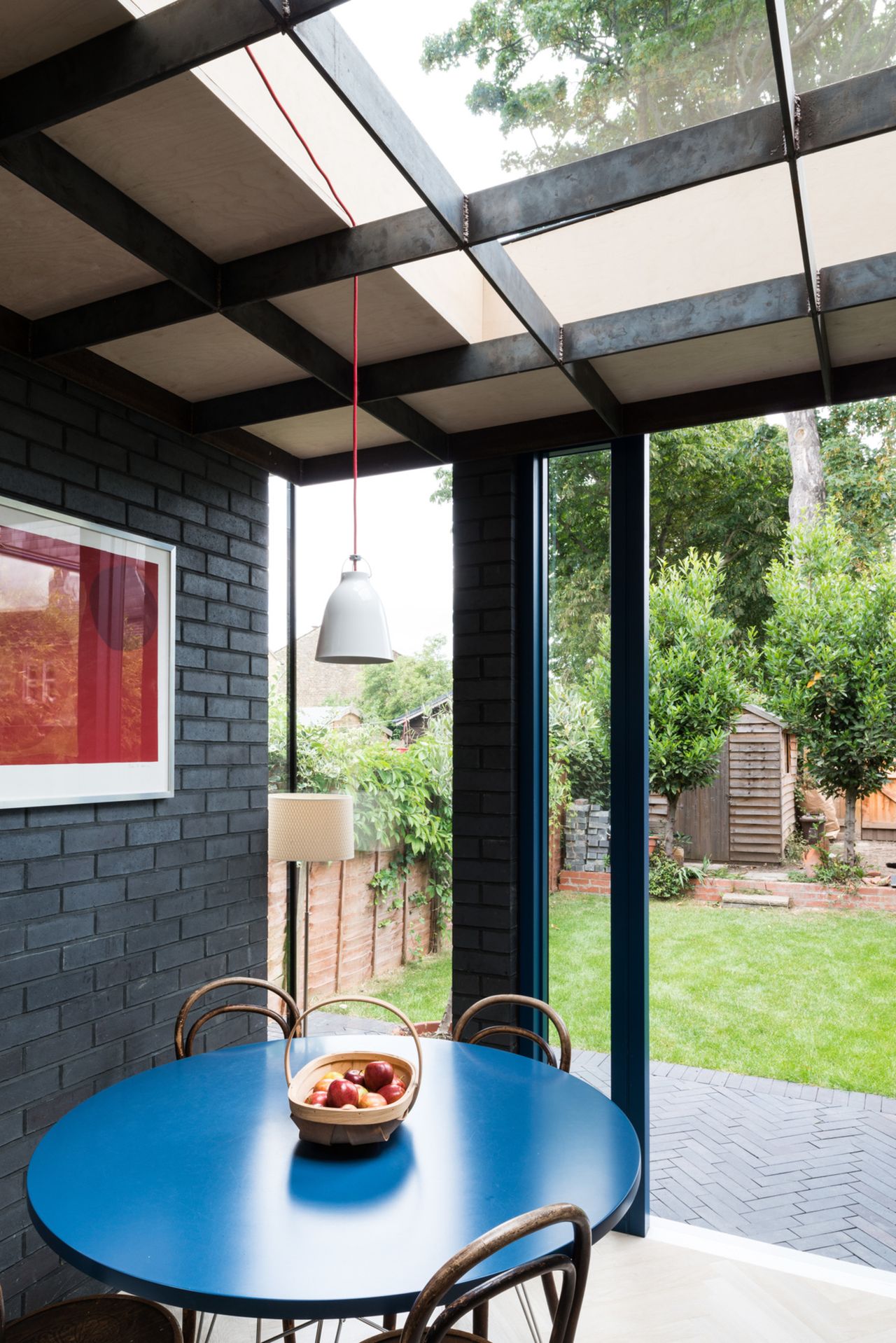 extension with glass roof, brick wall painted black, blue round table and pendant light hanging overhead, red artwork on the wall