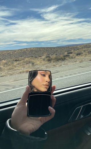 Lori Harvey takes a selfie in a compact mirror with a landscape behind her.