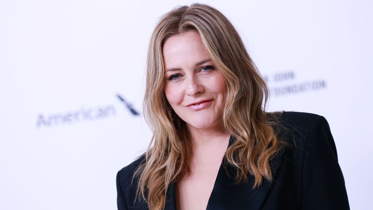 Alicia Silverstone wearing a black blazer, standing in front of a white background with black writing on it