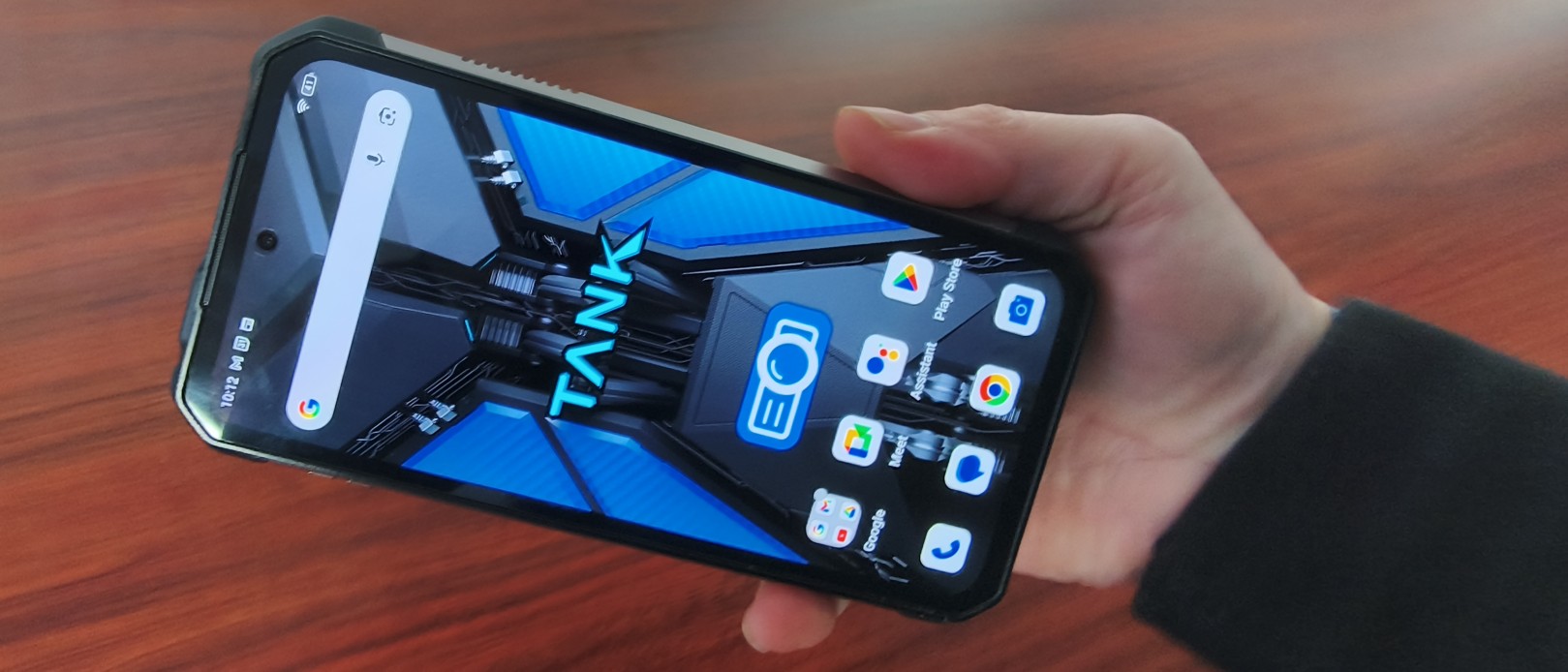 A person's hand holds the large, black Tank 4 Pro rugged smartphone at an angle, showing the blue and black "TANK" wallpaper on the display.