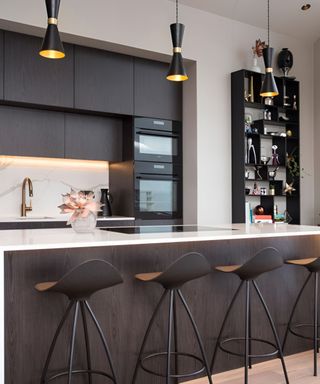 A kitchen with dark wooden cabinets with built-in ovens, a white kitchen island with a dark wooden back panel, three curved chairs in front of it and three black pendant lights above it
