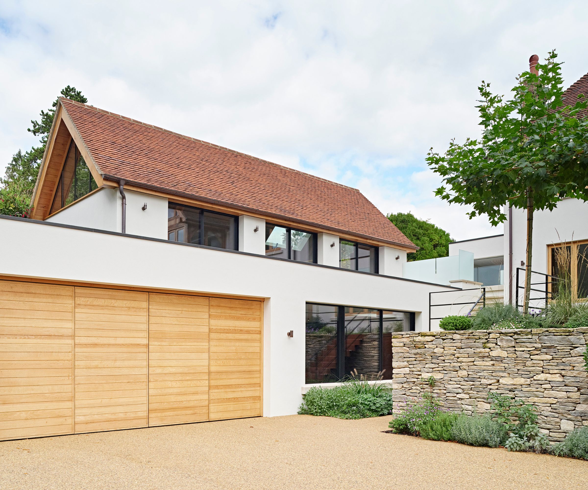 Modern home with a pitched clay tile roof and smooth white render, featuring large black-framed windows and light oak bifold garage doors. The driveway is a warm, golden gravel surface with a raised stone planter to one side