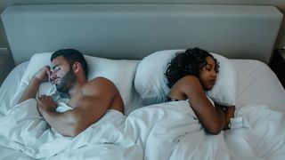 A couple asleep in bed under white bedsheets, lying back to back