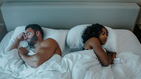 A couple asleep in bed under white bedsheets, lying back to back