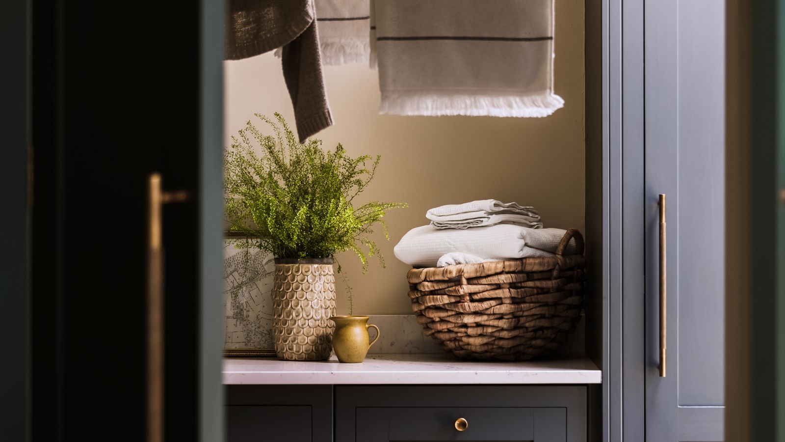 A glimpse through dark grey shaker cabinet doors with brass handles into a neatly styled alcove with a fern in a textured pot, wicker basket of folded towels and hanging linen.