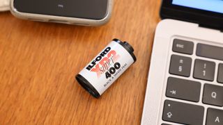 Ilford XP2 Super 35mm film canister on a wooden surface next to a MacBook