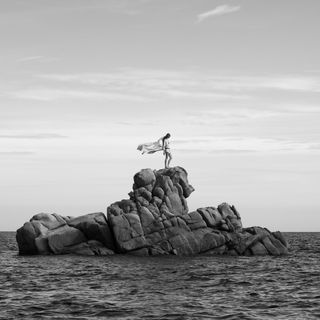 Black and white image of woman on rock with cape dress floating in breeze