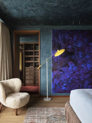 Image of a bedroom facing away from the bed. The walls are painted in a dark teal limewash and there is a giant indigo painting hanging on the wall. There is an off-white boucle arm chair and a floor lamp with a yellow shade in the corner.