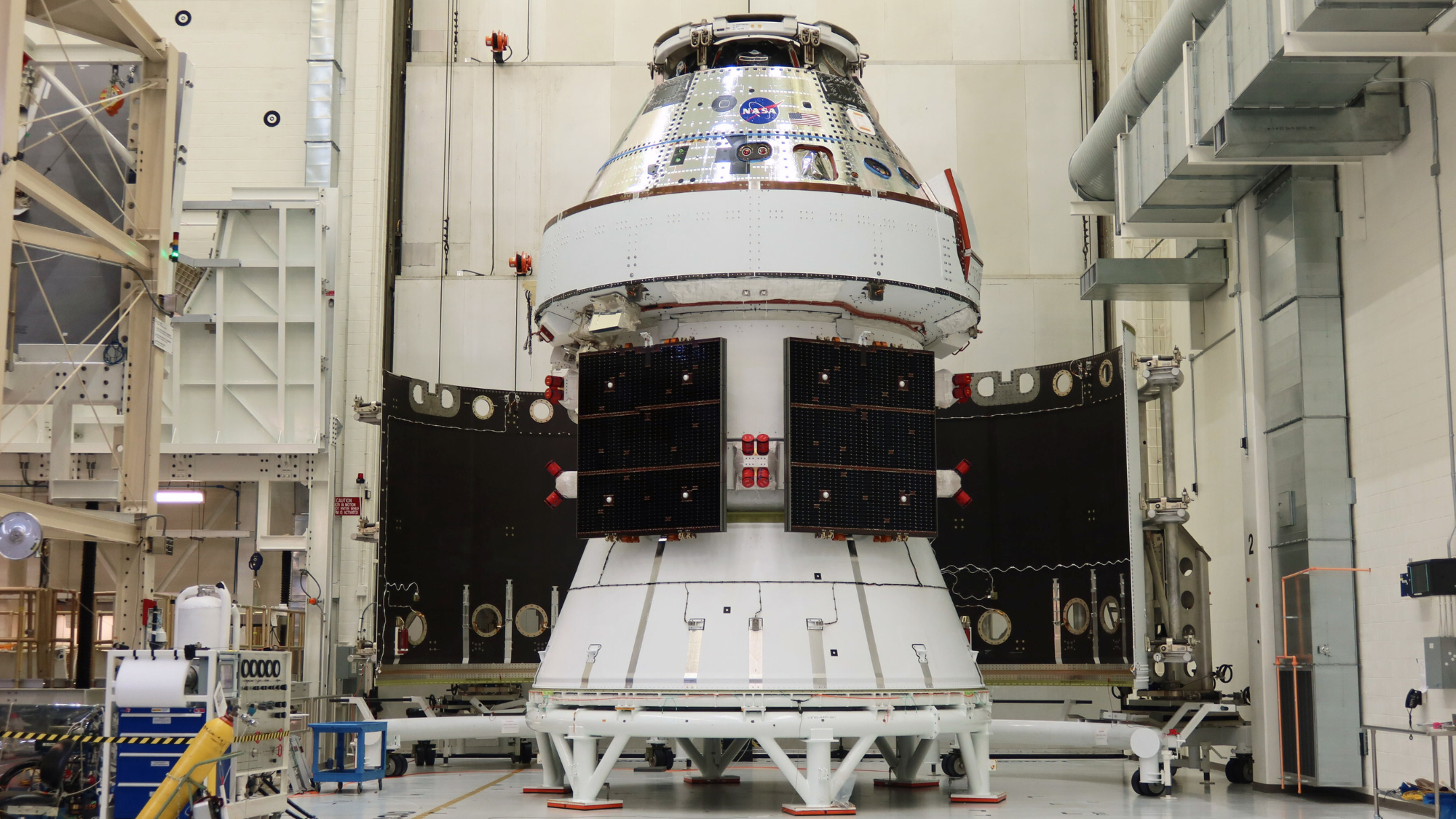 A silver space capsule sits within a white service module with folded square solar panels in a white warehous.