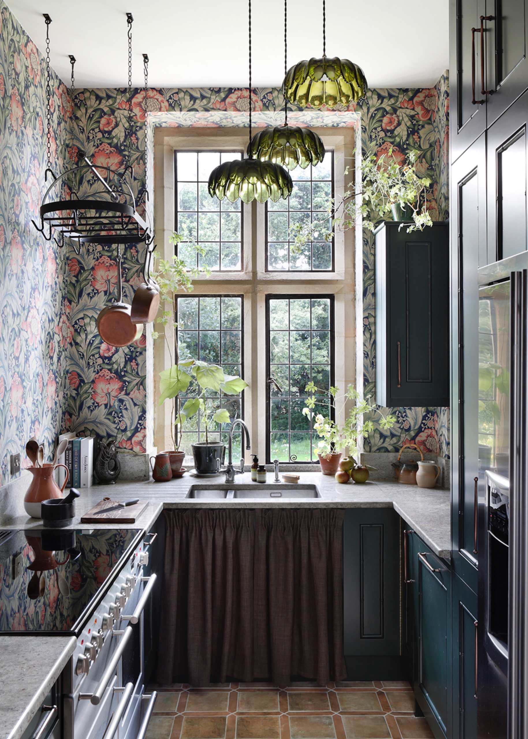 Studio Farra kitchen with deep green cabinets, botanical wallpaper, green glass pendant lights and pale stone countertops beneath a tall mullioned window.