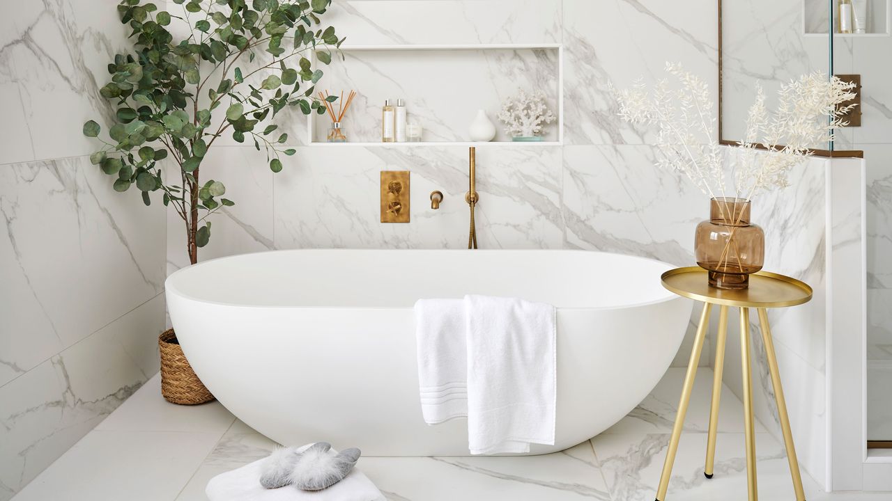 Freestanding bath in front of marble wall with gold side table and large house tree in corner in spa bathroom