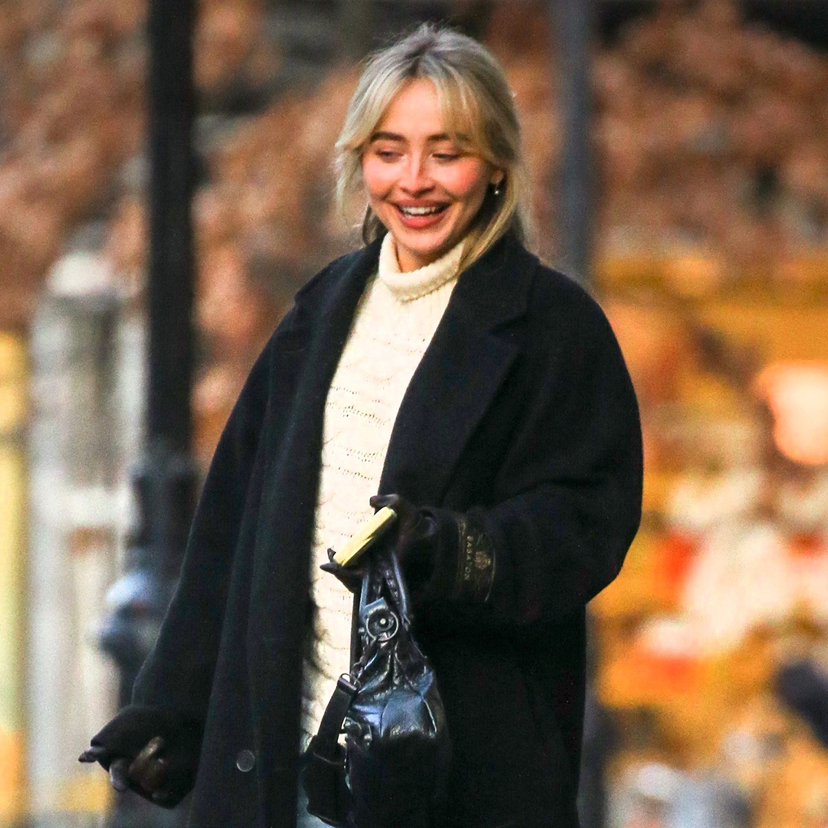Sabrina carpenter wears a black wool coat, ivory turtleneck and a black bag.