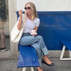 Model Lila Moss sits on a bench outside eating ice cream, wearing a white t-shirt, blue jeans, ballet flats and a white handbag.
