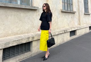 Eliza Huber at Fondazione Prada wearing a black sculpted Carven tunic with a yellow, satin Prada midi skirt, black wedge heels, and a black 1995 Prada bag.