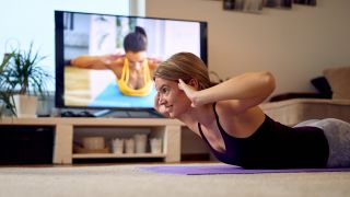 Woman performing dorsal raise in front of TV which shows trainer demonstrating dorsal raise