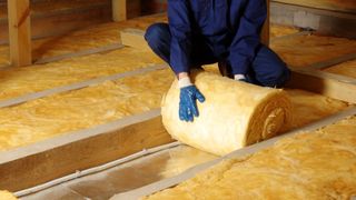 A man in a blue boiler suit unrolls insulation between rafters