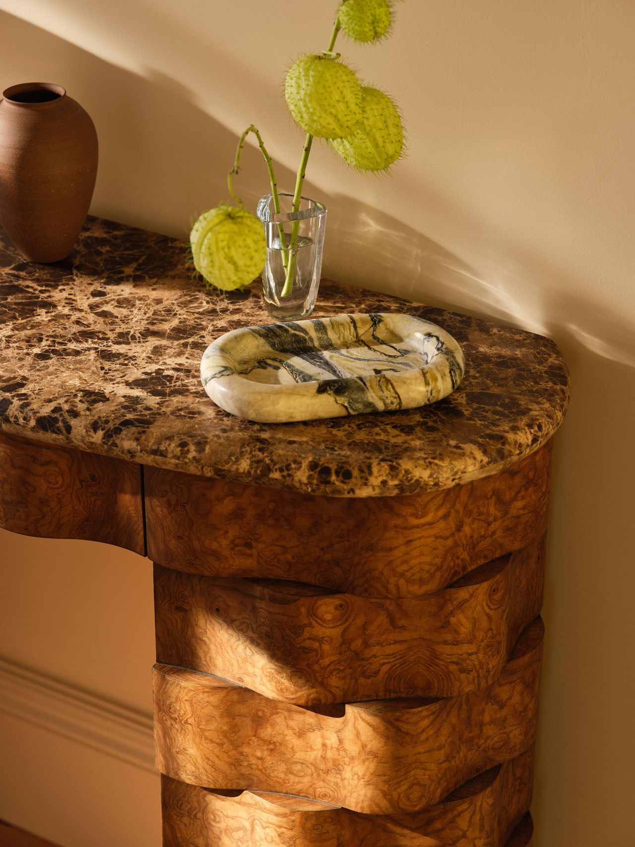 Close-up image of a wooden console table that is made in a woven design. The top of the table is a black and white granite. On top of the table is a decorative tray in a white and black marble material. There is a clear, small vase with lime-green bulbous flowers.