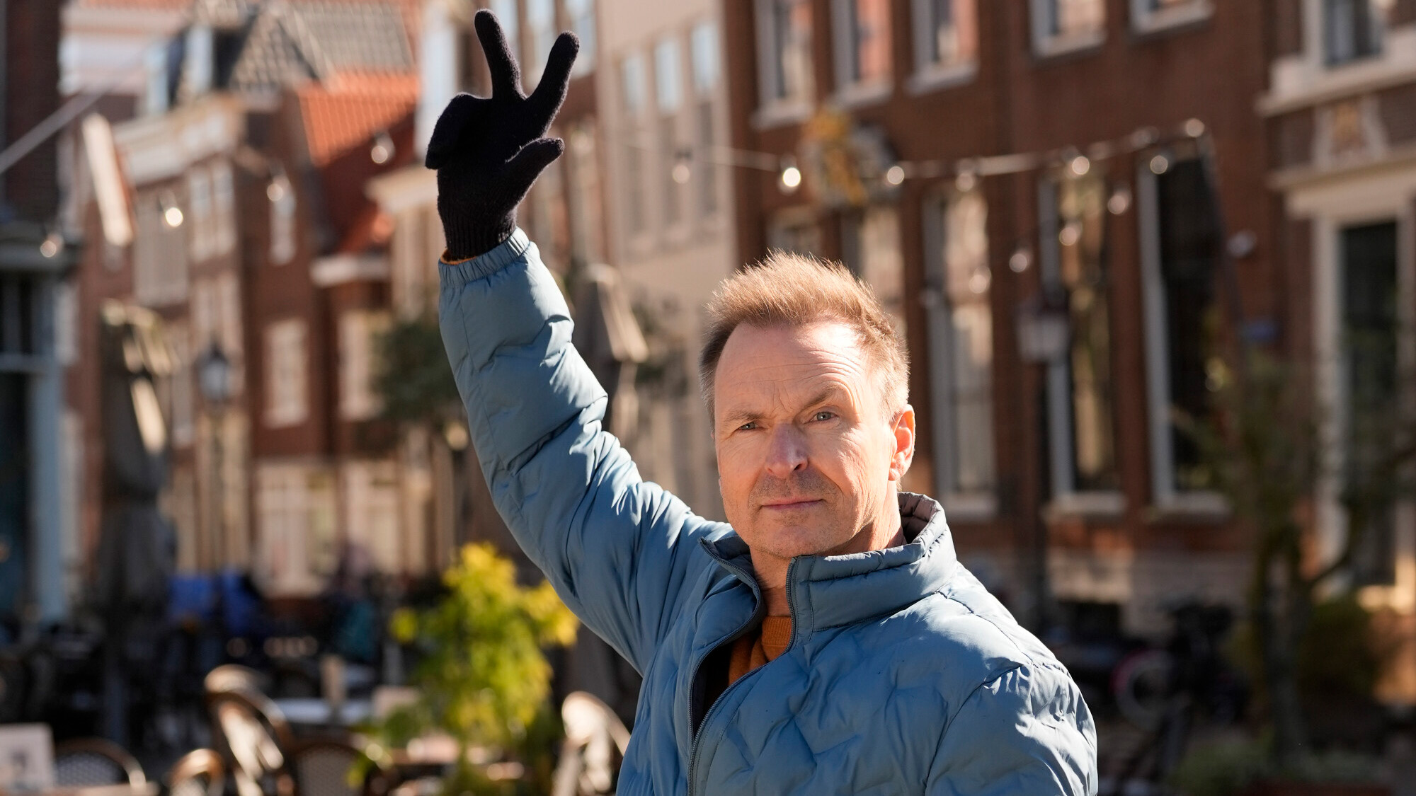 Phil raises two fingers in the air to greet a team in a blue coat and gloves on The Amazing Race.