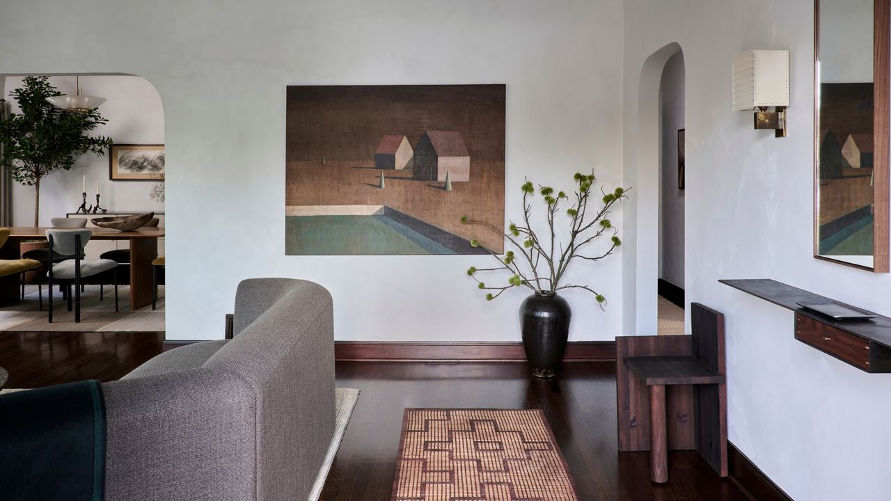 Image of a white living room with dark brown hardwood floors and trim. It is an open-concept room with a gray sofa and a runner rug in between the back of the sofa and the wall. There is a painting on the back wall and a large black vase on the floor