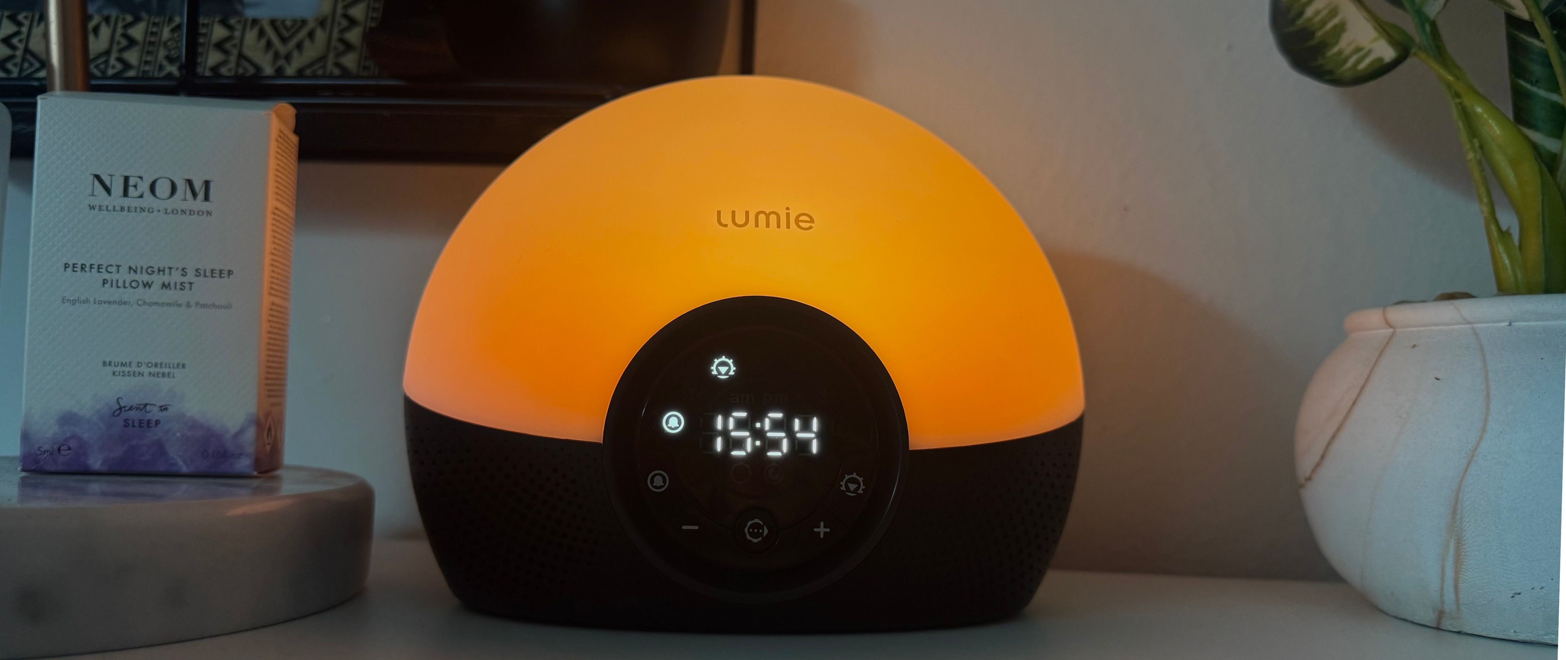 Lumie Bodyclock Glow 150 sunrise alarm clock on bedside table with NEOM pillow spray and plant 