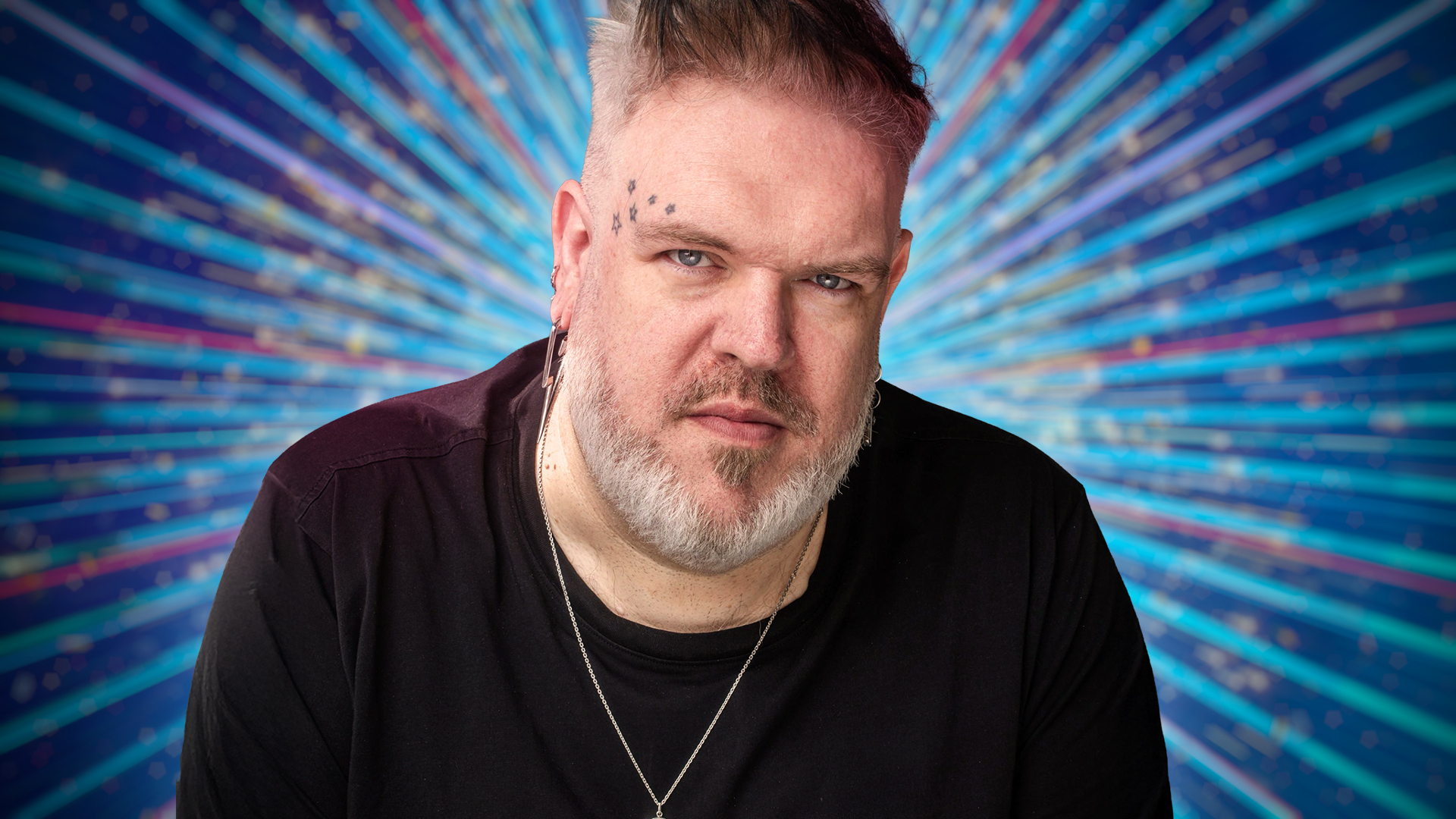 Kristian Nairn sitting with a blue sparkly background behind him.