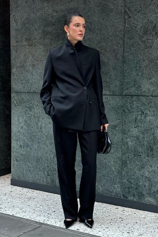 How to wear a suit is shown in a photo of a woman standing outside against a green marble background wearing gold earrings, a black blazers, a black handbag, black trousers, and black pointed pumps