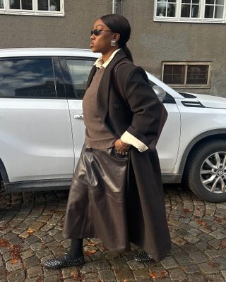 Woman wearing black coat, grey sweater, leather skirt, and white collared shirt.