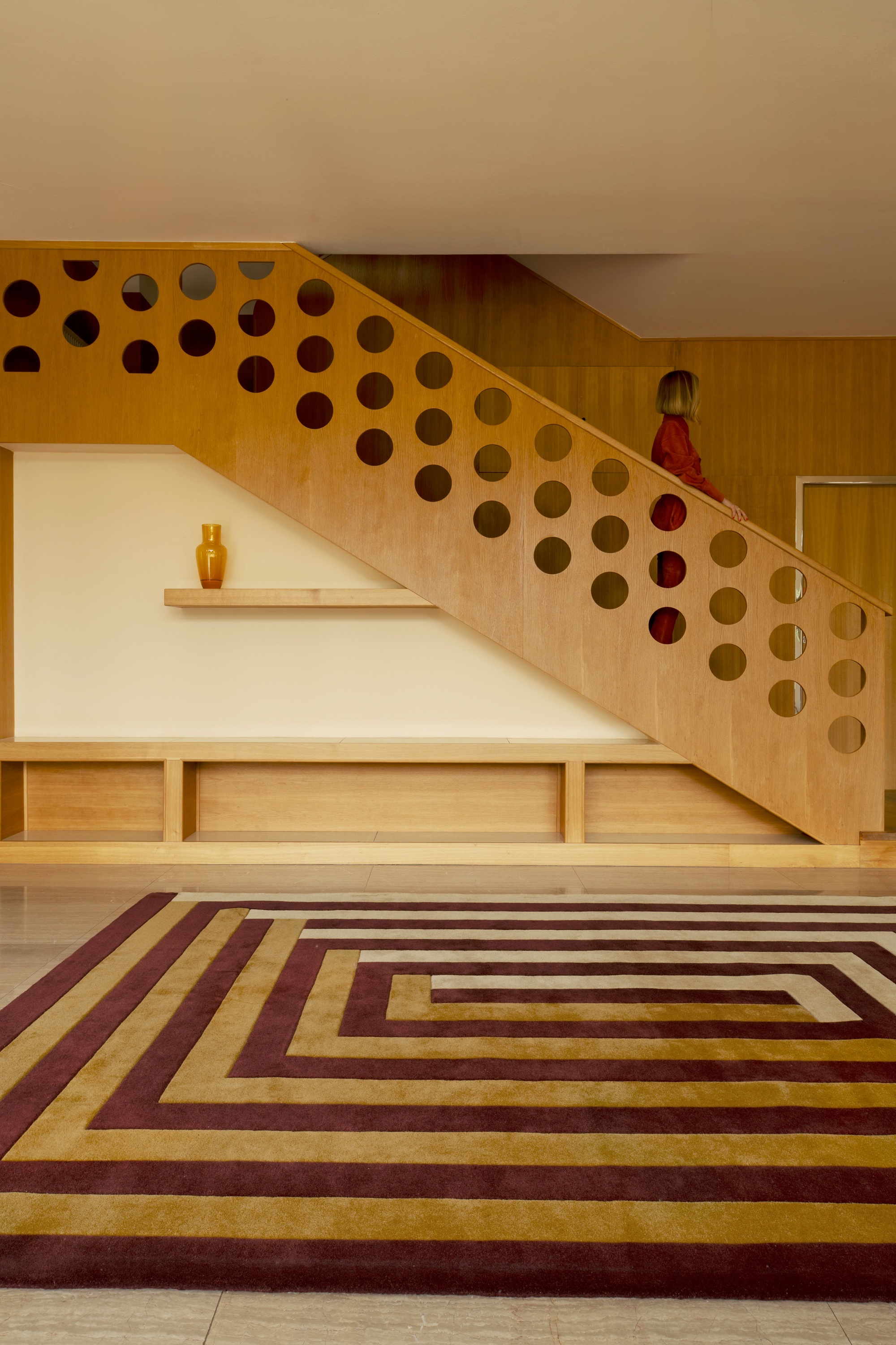 Image of an empty modern room with a perforated wooden stairwell with a woman in red walking down it. There is a large red, yellow, and cream striped rug on the ground.