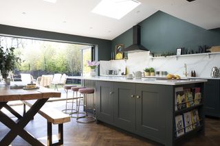 Open-plan kitchen diner with dark grey-green Shaker kitchen, parquet flooring, industrial-style dining table and benches, pink bar stools and bifold doors out to a decked seating area