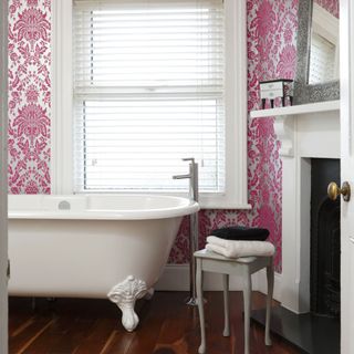 a bathroom with dark wooden floor and hot pink patterned walls, with a white freestanding clawfoot rolltop bathtub