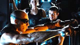 Still from the movie series The Chronicles of Riddick. Here we see Riddick (bald man wearing dark goggles) crouching down holding a blade in each hand. He is being watched by a man and woman in black armor.